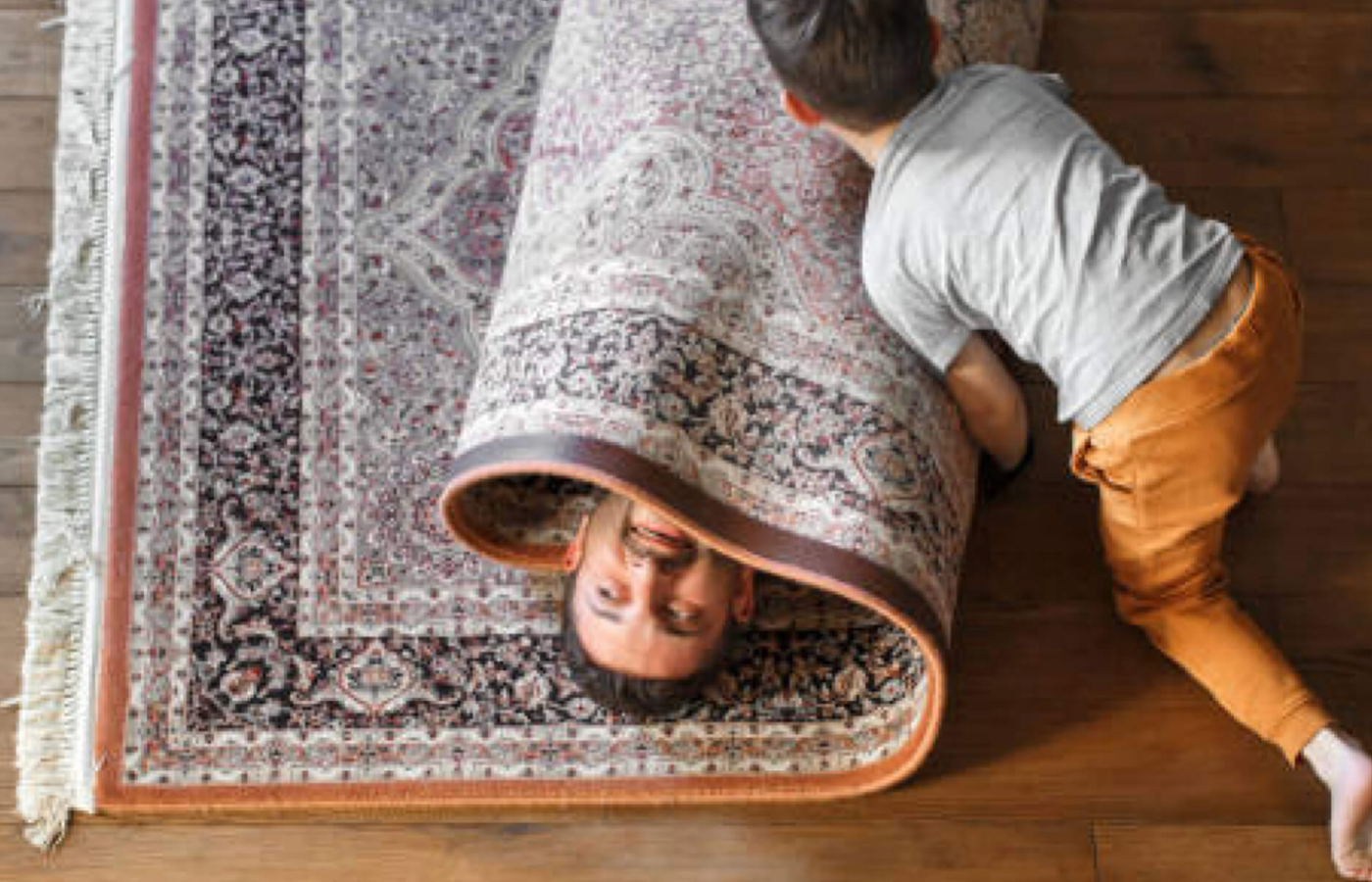 child rolling father up in a carpet playing on hardwood floors